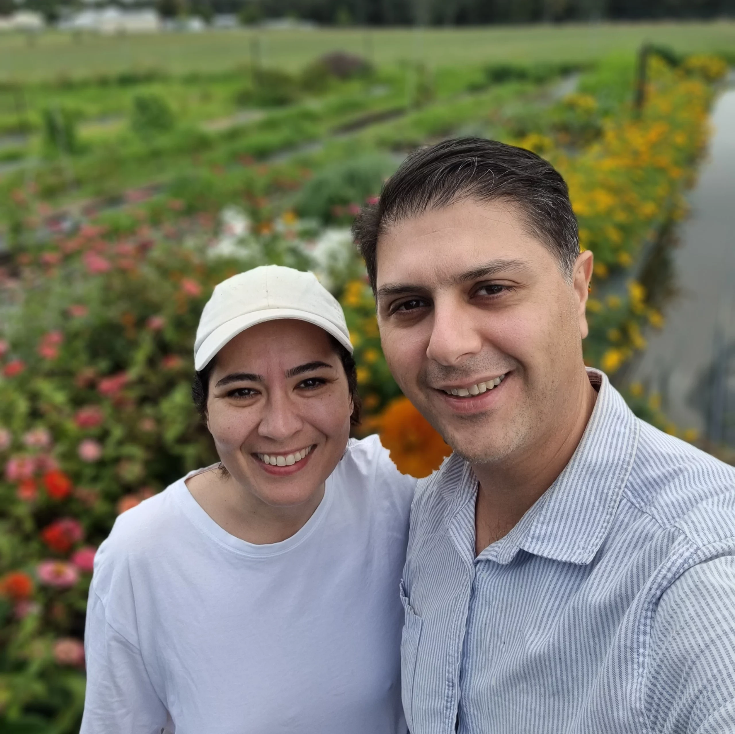 Azadeh and Harry, owners of Coomba Little Farm, smiling in front of a colorful flower garden.