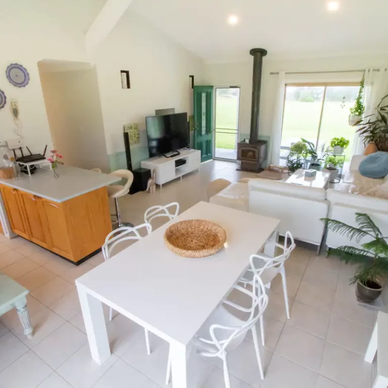 Bright open-plan living and dining area with white furniture, cozy fireplace, and natural light at Coomba Little Farm.