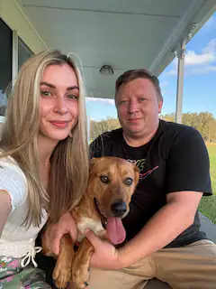 A happy couple sits on the porch of coomba little farm lakeside retreat, smiling warmly with their adorable brown dog sitting between them. The bright blue sky and lush greenery in the background highlight the peaceful, countryside setting of Coomba Little Farm, a perfect spot for a pet-friendly farm stay. Their dog looks content and relaxed, enjoying the outdoor serenity with its tongue playfully out, showing how the farm is a welcoming and cozy place for both people and pets alike.