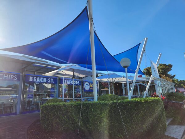 Exterior view of Beach Street Seafoods in Forster, featuring a vibrant blue shade sail, neatly trimmed hedges, and outdoor seating, creating a welcoming and relaxed dining atmosphere.