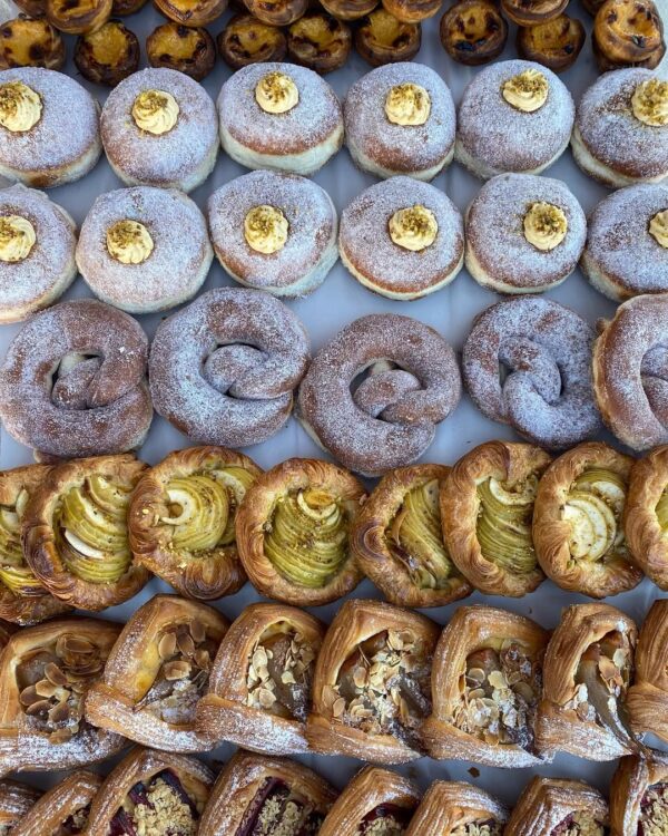 An array of freshly baked pastries from Palms Artisan Bakery in Forster, featuring cream-filled donuts, sugar-dusted pretzels, apple danishes, and almond-topped delights.