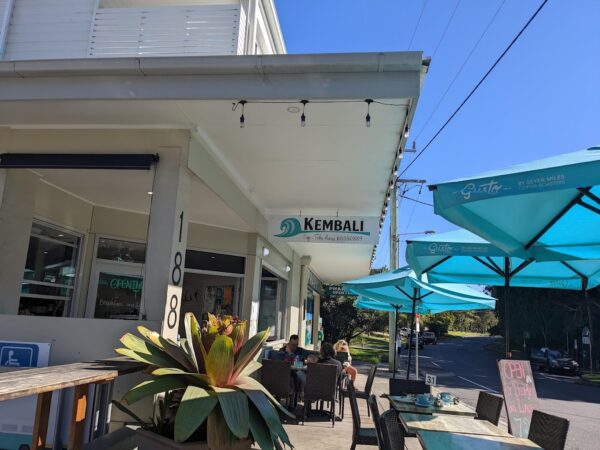 Street view of Kembali Café at 188 Boomerang Drive, Blueys Beach, featuring outdoor seating with turquoise umbrellas, lush plants, and a bright sunny atmosphere.