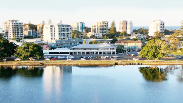 A scenic view of the Lakes & Ocean Hotel in Forster, situated along the waterfront with a backdrop of modern buildings, lush greenery, and calm reflective waters.