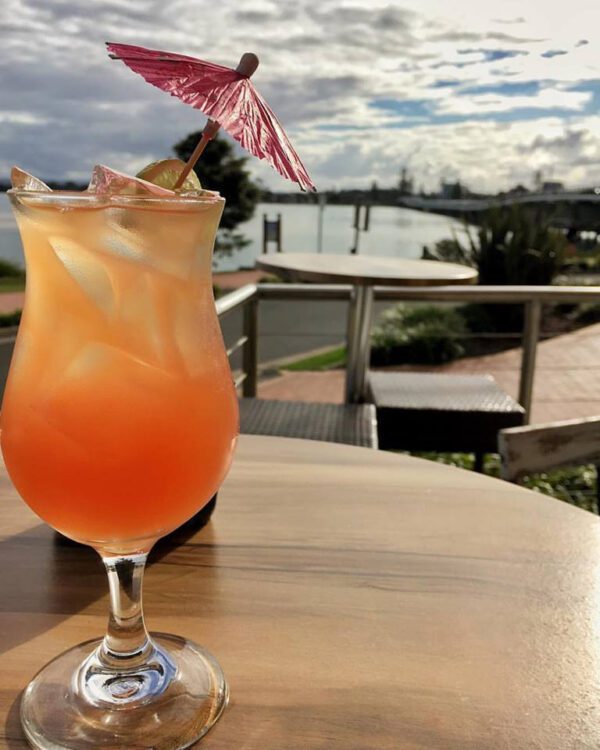 Tropical cocktail with a pink umbrella served at Spice Monkey in Forster, showcasing a stunning view of Wallis Lake and its serene surroundings.